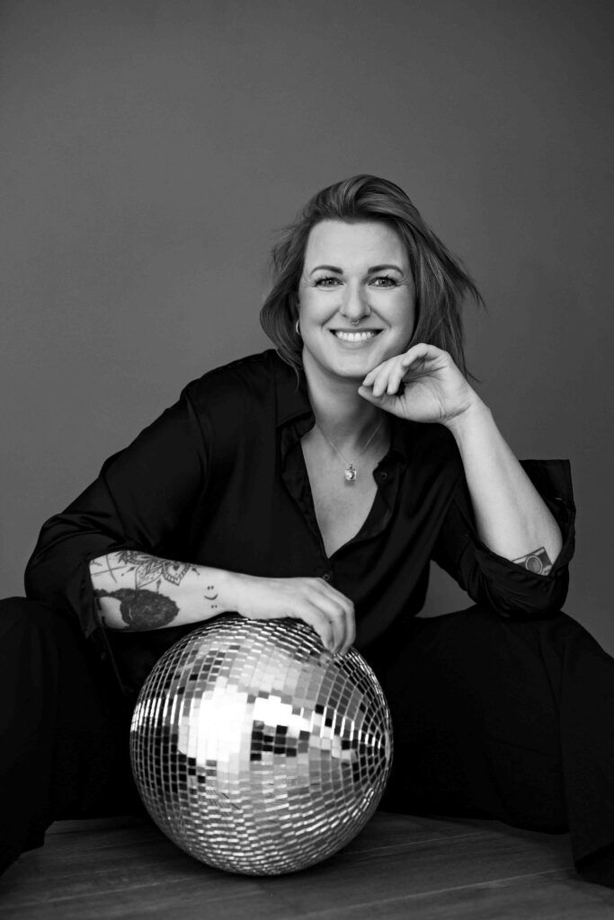 Foto zeigt mich als Fotografin in schwarzer Kleidung. Sitze auf dem Boden mit einer Discokugel vor mir und einem herzlichen, offenen Lächeln