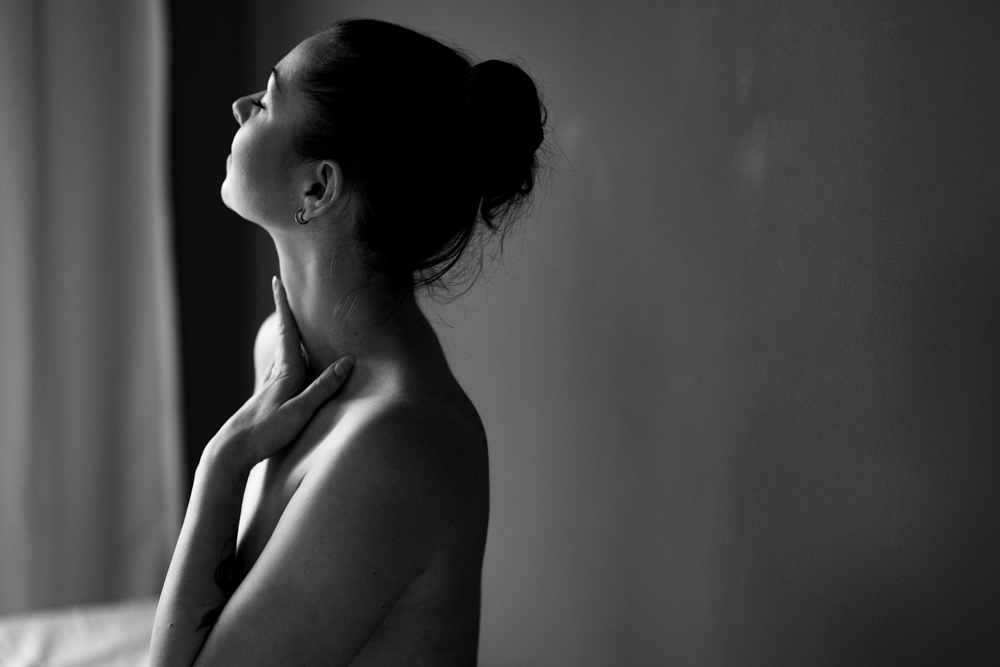 Frau wird seitlich fotografiert. Ihre Hand gleitet über ihren Hals. Sie ist nackt aber das Bild ist sehr ästhetisch