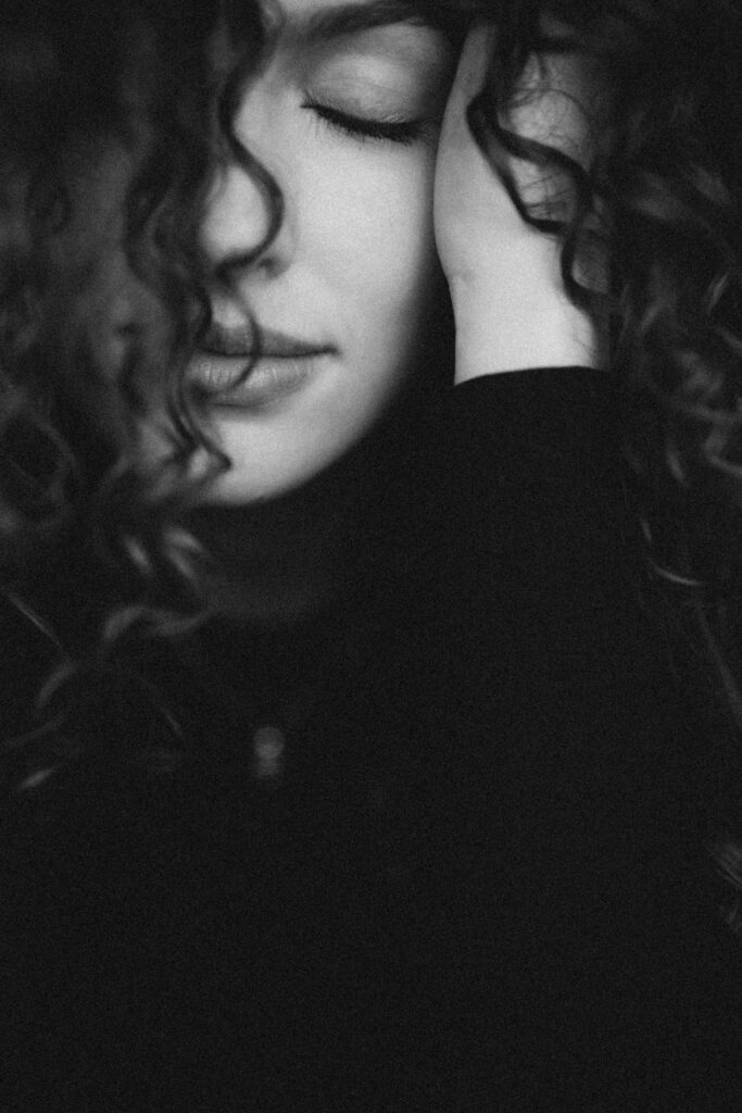 Sinnliches Close Up Porträt in schwarz weiß von einer Frau mit vielen Locken, die ihr über das Gesicht hängen. Sie hat geschlossene Augen